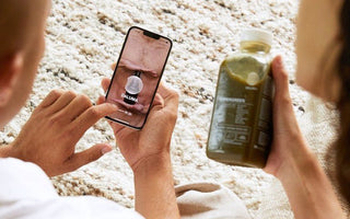 Person hält ein Smartphone mit geöffneter DALUMA App, daneben hält eine zweite Person eine Flasche Supergreen Saft über einem hellen Wollteppich