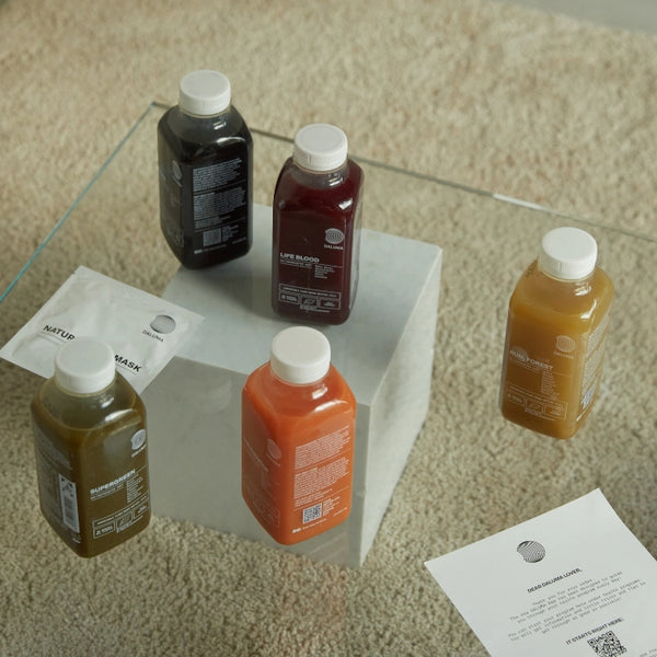 Vier Flaschen DALUMA CLEANSE DAY, jede in einer anderen Farbe, sind auf und um einen weißen Block auf einem beigen Teppich angeordnet. In der Nähe liegen zwei Stücke Papier - eines gefaltet, eines flach - und vervollständigen die sanft beleuchtete Szene.