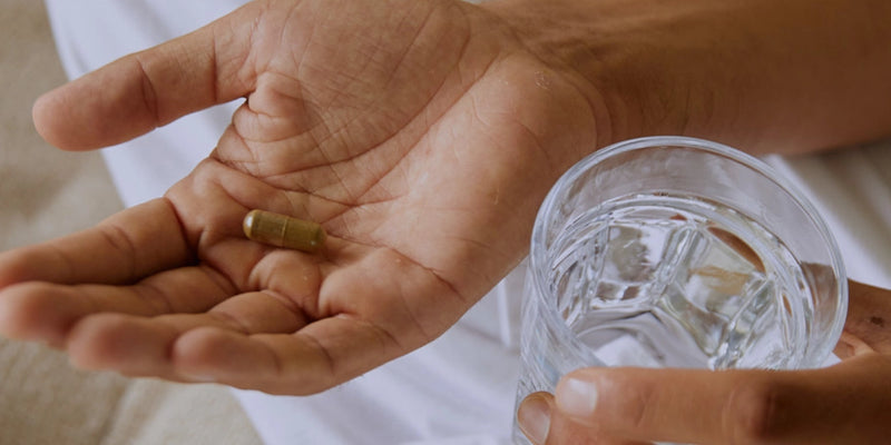 Eine Person hält eine braune Kapsel in der einen Hand und ein klares Glas Wasser in der anderen und bereitet sich auf die Einnahme des Medikaments vor.
