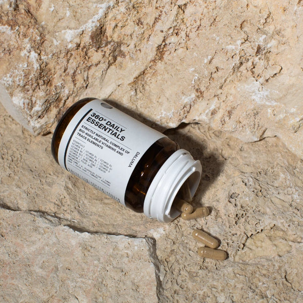 Eine Flasche mit der Aufschrift 360° Daily Essentials liegt auf einer strukturierten Steinfläche, daneben liegen mehrere beigefarbene Kapseln.