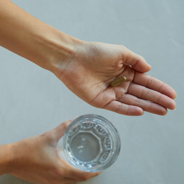 Eine Person hält ein Glas Wasser und eine einzelne grüne DALUMA 360 DAILY ESSENTIALS SUPPLEMENT Kapsel vor einem hellen Hintergrund, der die moderne pflanzliche Ernährung und Mikronährstoffe hervorhebt.