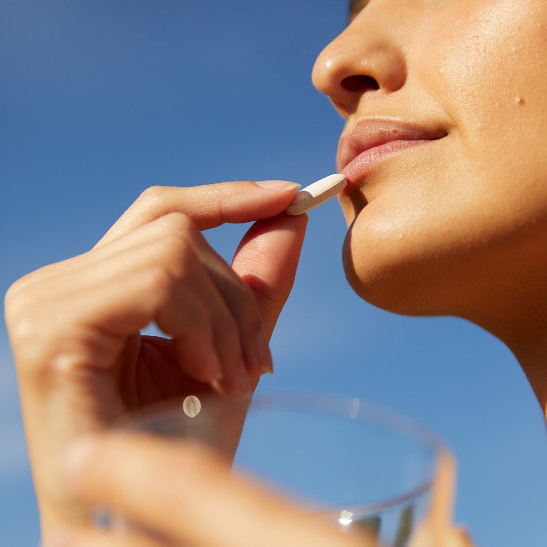 Eine Person hält eine weiße Pille an ihre Lippen und ein Glas Wasser und bereitet sich auf die Einnahme von SIX ELEMENTS SKIN KOLLAGEN KAPSELN von DALUMA vor. Im Hintergrund ist ein klarer blauer Himmel zu sehen.