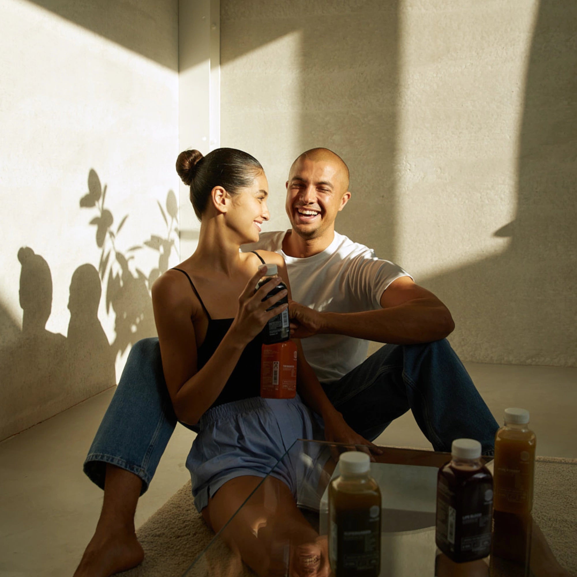 Ein lächelndes Paar sitzt auf dem Boden im Sonnenlicht, hält DALUMA BETTER GUT SAFTKUR 7 TAGE Säfte in der Hand und schaut sich an. Drei weitere Flaschen stehen auf einem Glastisch in der Nähe, während sich Blätterschatten an der Wand bewegen - ideal für Ihre Saftkur.