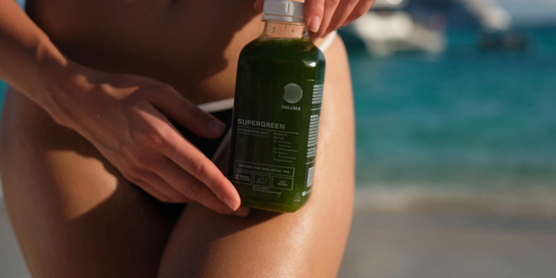 Eine Person im Badeanzug hält eine grüne Saftflasche mit der Aufschrift Supergreen in der Nähe ihres Beins an einem sonnigen Strand, mit blauem Wasser und einem verschwommenen Boot im Hintergrund.
