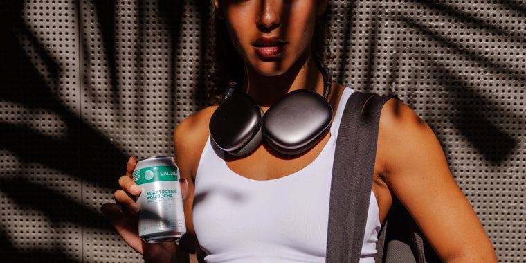 Eine Person in einem weißen Tank-Top mit großen Kopfhörern um den Hals hält eine Dose mit sprudelndem Wasser. Die Schatten der nahen Palmenblätter werfen sich auf ihr Gesicht und die perforierte Metallwand hinter ihr.