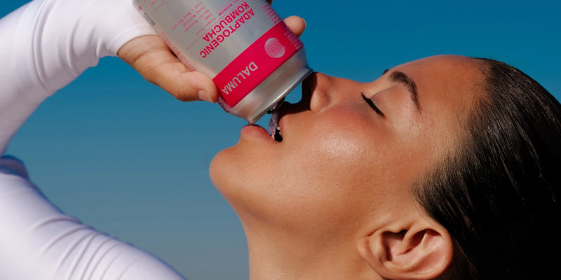 Eine Person mit geglättetem Haar und leuchtender Haut trinkt aus einer Dose mit der Aufschrift DALUMA ADAPTOGENIC KOMBUCHA vor einem strahlend blauen Himmel und trägt ein weißes langärmeliges Oberteil.