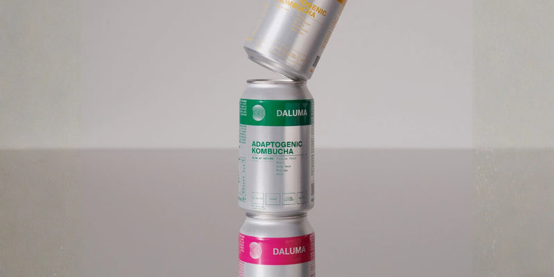 Drei Dosen Daluma-Kombucha sind senkrecht gestapelt, wobei die oberste Dose gekippt ist. Die Dosen haben Etiketten in verschiedenen Farben: Gold, Grün und Rosa. Der Hintergrund ist schlicht und minimalistisch.