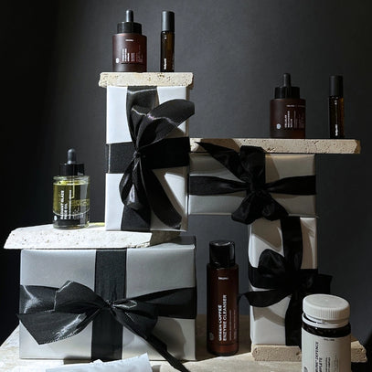 Mehrere Hautpflegeflaschen und Tropfflaschen werden auf und um gestapelte, mit schwarzem Band verpackte Schachteln vor einem dunklen Hintergrund und bei dramatischer Beleuchtung ausgestellt.