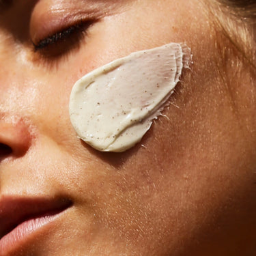 Nahaufnahme des Gesichts einer Person mit geschlossenen Augen, auf deren Wange ein dicker Klecks DALUMA DEEP HYDRATION FEUCHTIGKEITSMASKE mit natürlichen Zutaten und Shea Butter aufgetragen ist; die glatte Haut leuchtet im Sonnenlicht.
