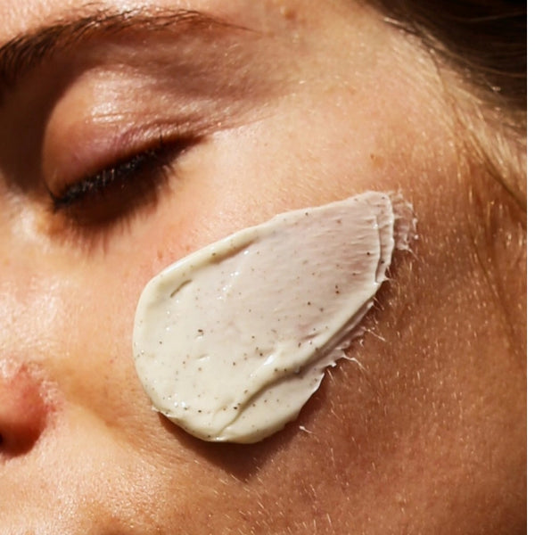 Eine Nahaufnahme des Gesichts einer Person mit geschlossenen Augen, auf deren Wange ein dickflüssiges, cremiges Hautpflegeprodukt mit kleinen Peelingpartikeln aufgetragen wurde.