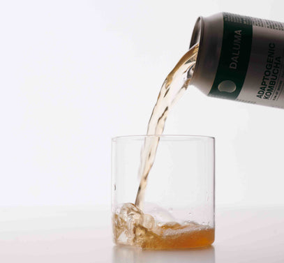Eine Dose mit der Aufschrift Adaptogenic Kombucha wird gekippt und gießt eine hellbraune Flüssigkeit in ein klares Glas mit einem Spritzer. Der Hintergrund ist weiß und minimalistisch.
