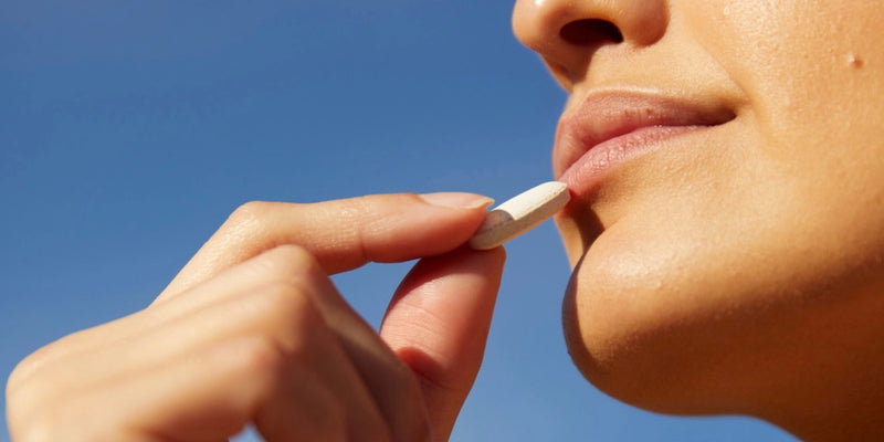 Nahaufnahme einer Person, die eine weiße Pille vor dem Hintergrund eines klaren blauen Himmels an die Lippen hält und sich auf die Einnahme des Medikaments vorbereitet.