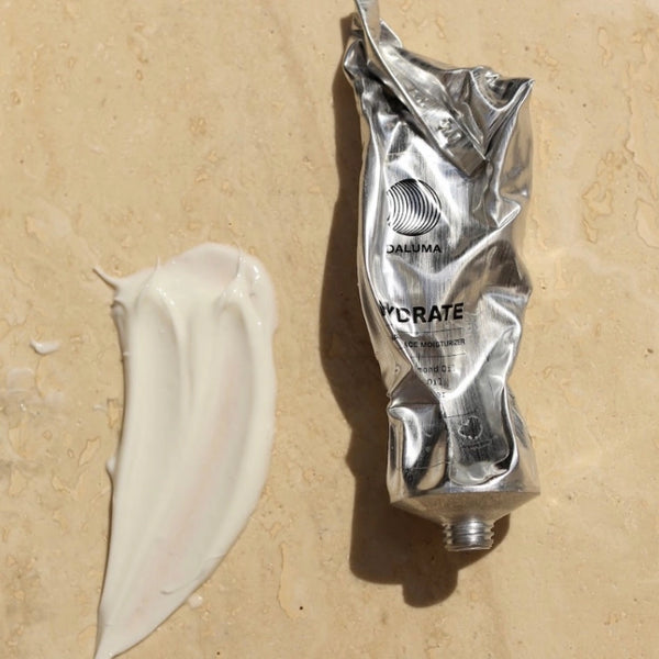 Eine zerknitterte Metalltube mit der Aufschrift DAIJUMA HYCRATE liegt auf einer beigen Fläche neben einem dicken Klecks weißer Creme.