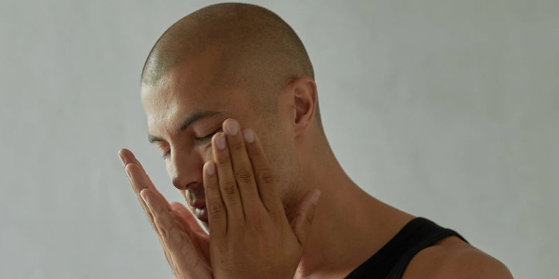 Ein Mann mit rasiertem Kopf und schwarzem Tank-Top hält sich die Hände vors Gesicht, die Augen geschlossen, vor einem schlichten hellgrauen Hintergrund.