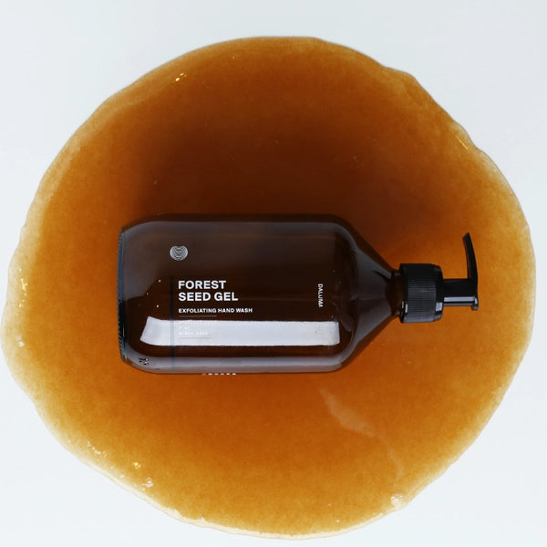Eine braune Pumpflasche mit der Aufschrift Forest Seed Gel Exfoliating Hand Wash liegt auf einer kreisförmigen Lache aus bernsteinfarbenem Gel vor einem weißen Hintergrund.
