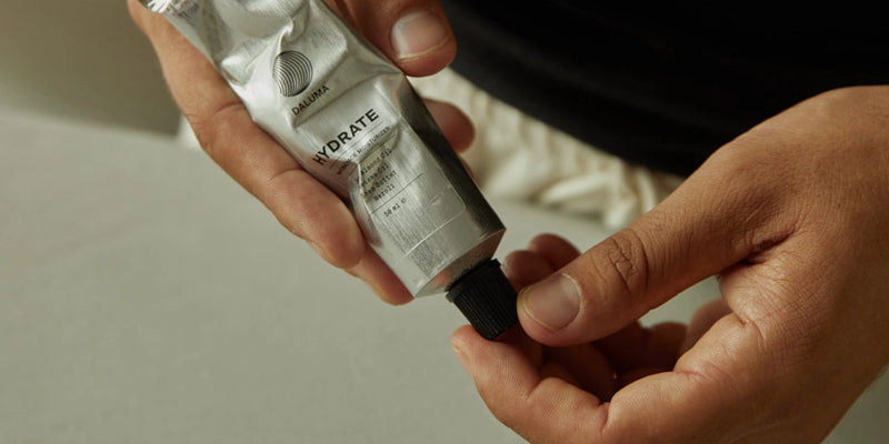 Eine Person hält eine Metalltube mit der Aufschrift HYDRATE" in der Hand und schraubt mit einer Hand den schwarzen Deckel ab, um das Produkt, möglicherweise eine Lotion oder Creme, zu benutzen.