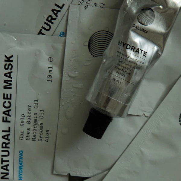 Eine Nahaufnahme von Hautpflegeprodukten, darunter Maskenpackungen mit der Aufschrift Natural Face Mask und Inhaltsstoffen sowie eine Metalltube mit der Aufschrift Hydrate, die alle Wassertropfen auf der Oberfläche aufweisen.