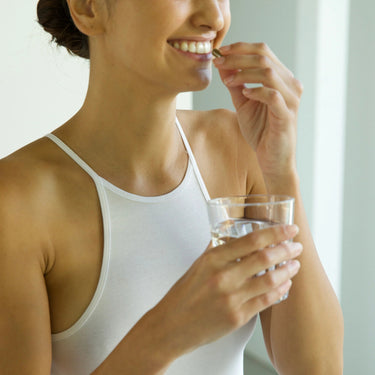 Eine Frau in einem weißen Tank-Top lächelt, während sie ein Glas Wasser hält und DALUMA IMMUN DEFENCE COMPLETE einnimmt, um ihr Immunsystem zu stärken und die natürlichen Abwehrkräfte ihres Körpers zu unterstützen.