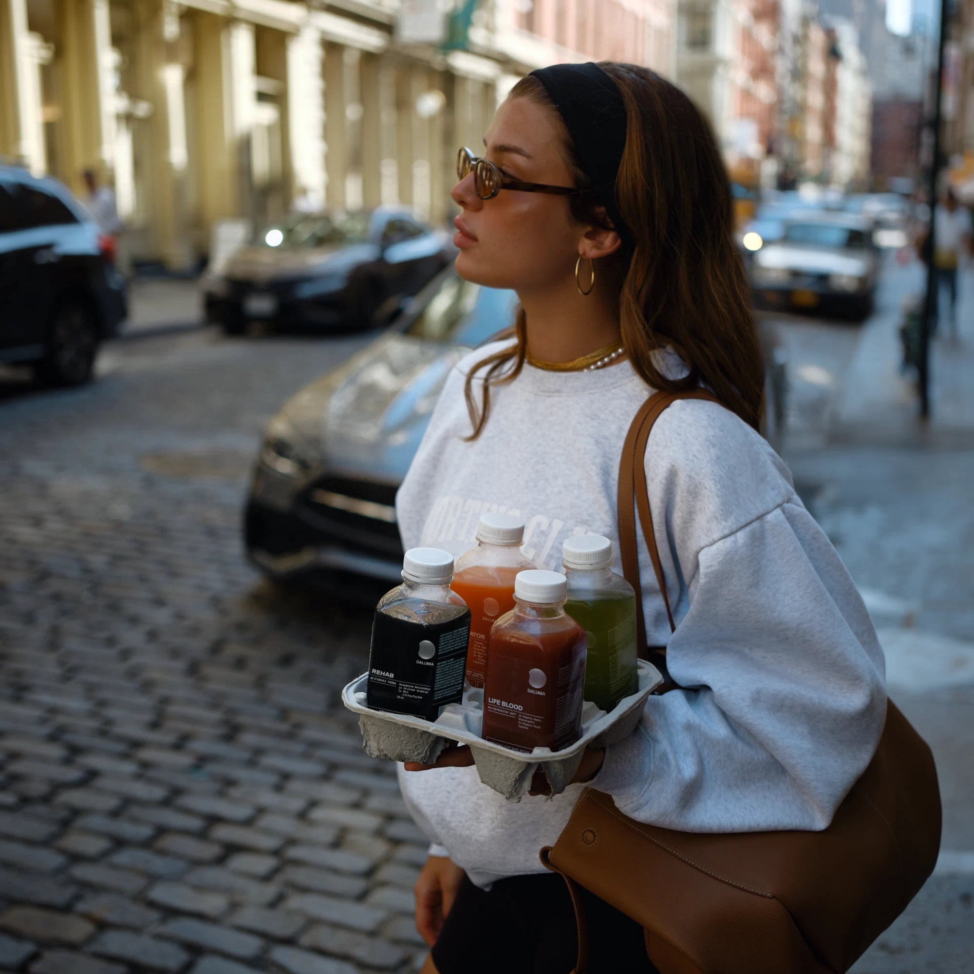 Eine Frau mit Sonnenbrille und Sweatshirt geht eine sonnige, kopfsteingepflasterte Straße entlang und trägt die Flaschen BESSERE SAFTKUR - 7 TAGE von DALUMA. Autos und Gebäude säumen den Hintergrund.
