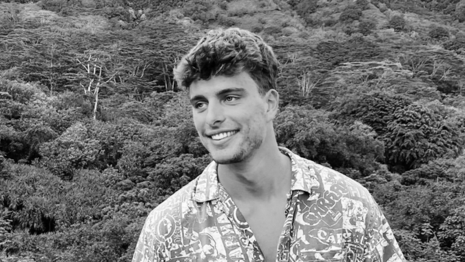 Ein junger Mann mit kurzen Haaren und einem gemusterten Hemd lächelt, während er im Freien steht, mit dichten, belaubten Bäumen und Blättern im Hintergrund. Das Foto ist in schwarz-weiß gehalten.