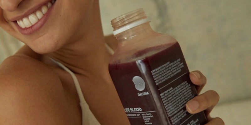 Eine lächelnde Person, die eine Flasche dunkelroten Saft mit einem teilweise sichtbaren Etikett in der Hand hält und ein helles Tanktop trägt.