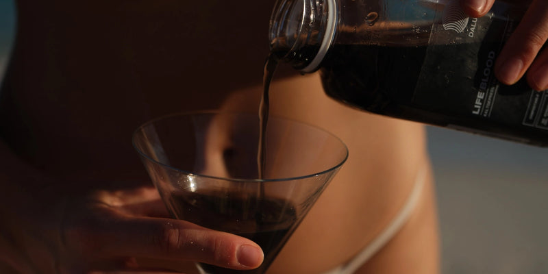 Eine Person im Bikini hält ein Martini-Glas und gießt eine dunkle Flüssigkeit aus einer Plastikflasche ein. Der Hintergrund ist leicht unscharf und konzentriert sich auf die Hände und den Einschenkvorgang.