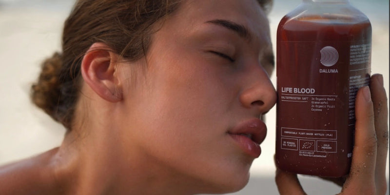 Eine Frau mit geschlossenen Augen hält eine Flasche mit der Aufschrift LIFE BLOOD nahe an ihr Gesicht. Die Flasche enthält eine rote Flüssigkeit und ist mit Markenzeichen und Produktinformationen versehen. Der Hintergrund ist sanft verschwommen.