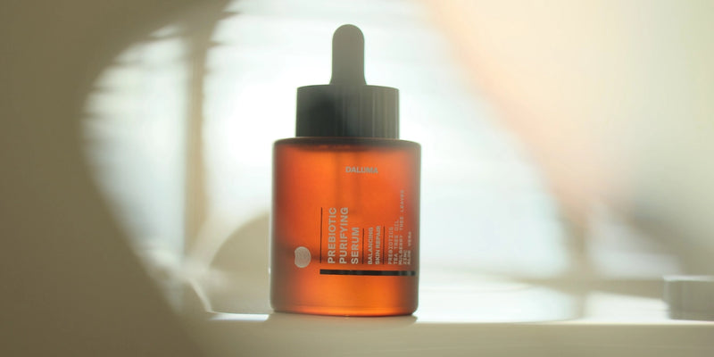 Eine braune Glastropfflasche mit der Aufschrift Prebiotic Serum steht auf einer weißen Fläche mit weichem, natürlichem Licht und verschwommenen Schatten im Hintergrund.