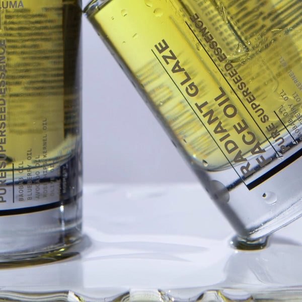 Zwei durchsichtige Flaschen mit gelb gefärbtem Gesichtsöl, die mit "Radiant Glaze" und "Pure Supersede Essence" beschriftet sind, ruhen auf einer spiegelnden Oberfläche und erzeugen einen Glanzeffekt mit Öl- und Flüssigkeitsreflexen.