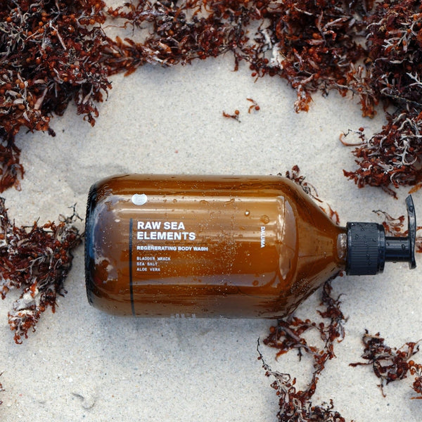 Eine braune Pumpflasche mit der Aufschrift Raw Sea Elements Regenerating Body Wash liegt auf dem nassen Sand, umgeben von verstreuten rötlich-braunen Algenstücken.