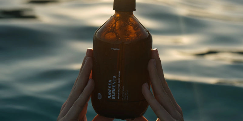 Ein Paar Hände hält eine braune, wassergetränkte Flasche mit der Aufschrift RAW SEA ELEMENTS über dem plätschernden blauen Wasser, auf dessen Oberfläche sich das Sonnenlicht spiegelt.