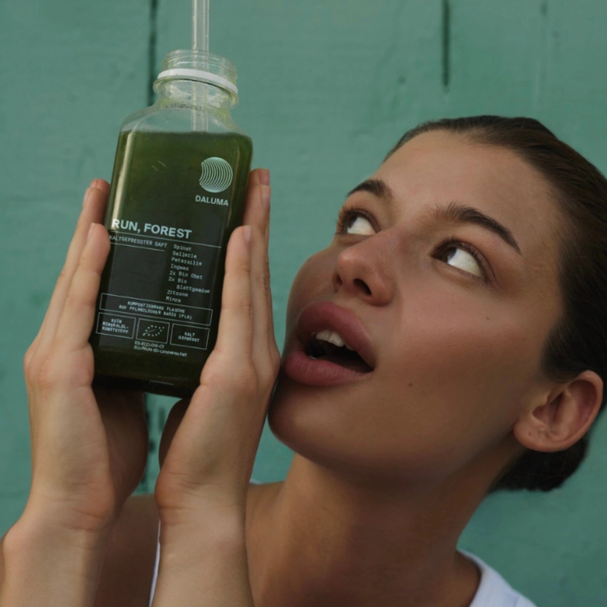 Eine Frau hält eine Flasche DALUMA RUN, FOREST GREEN JUICE 6 PACK in der Hand und blickt sie überrascht an. Der blaugrüne Hintergrund hebt den Saft hervor, der mit dem Kaltpressverfahren hergestellt wurde.