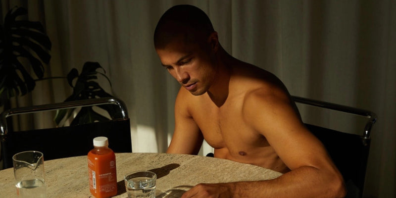 Ein Mann ohne Hemd sitzt mit nachdenklicher Miene an einem runden Tisch. Auf dem Tisch stehen eine Flasche mit Saft, ein Glas Wasser und ein weiteres Glas. Der Hintergrund ist sanft beleuchtet, ein Vorhang und eine Blattpflanze sind zu sehen.