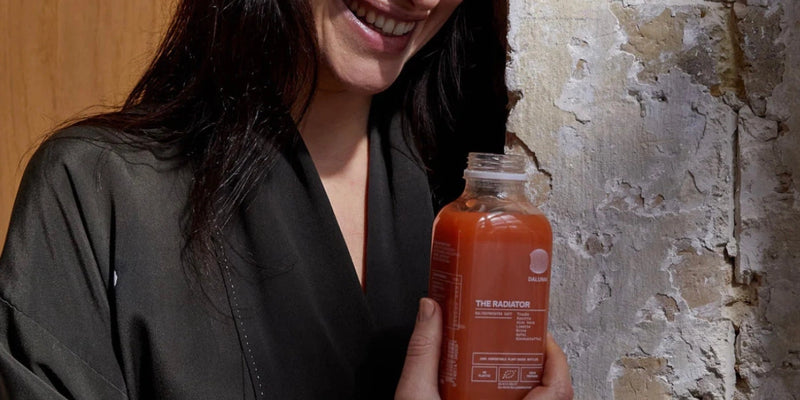 Eine Person in einem dunklen Gewand lächelt, während sie eine Flasche mit orange-roter Flüssigkeit mit der Aufschrift THE RADIATOR vor einer strukturierten, verwitterten Wand hält.