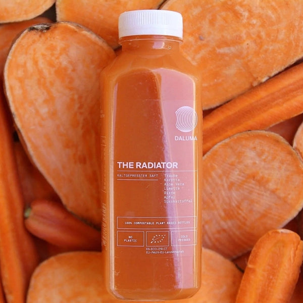 Eine Flasche des Daluma The Radiator-Saftes steht aufrecht, umgeben von herzförmigen Scheiben aus orangefarbener Süßkartoffel. Das Etikett listet die Zutaten auf und hebt die umweltfreundliche, pflanzliche Verpackung hervor.