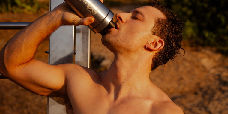 Ein Mann ohne Hemd trinkt aus einer Wasserflasche aus rostfreiem Stahl im Freien in warmem Sonnenlicht, mit einem unscharfen natürlichen Hintergrund hinter ihm.