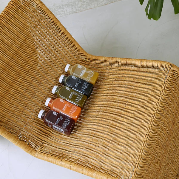 Vier kleine Saftflaschen in verschiedenen Farben (lila, orange, grün und gelb) sind in einer Reihe auf einem geflochtenen Rattanstuhl mit gebogener Rückenlehne angeordnet. In der oberen rechten Ecke ist teilweise eine grüne Pflanze zu sehen.