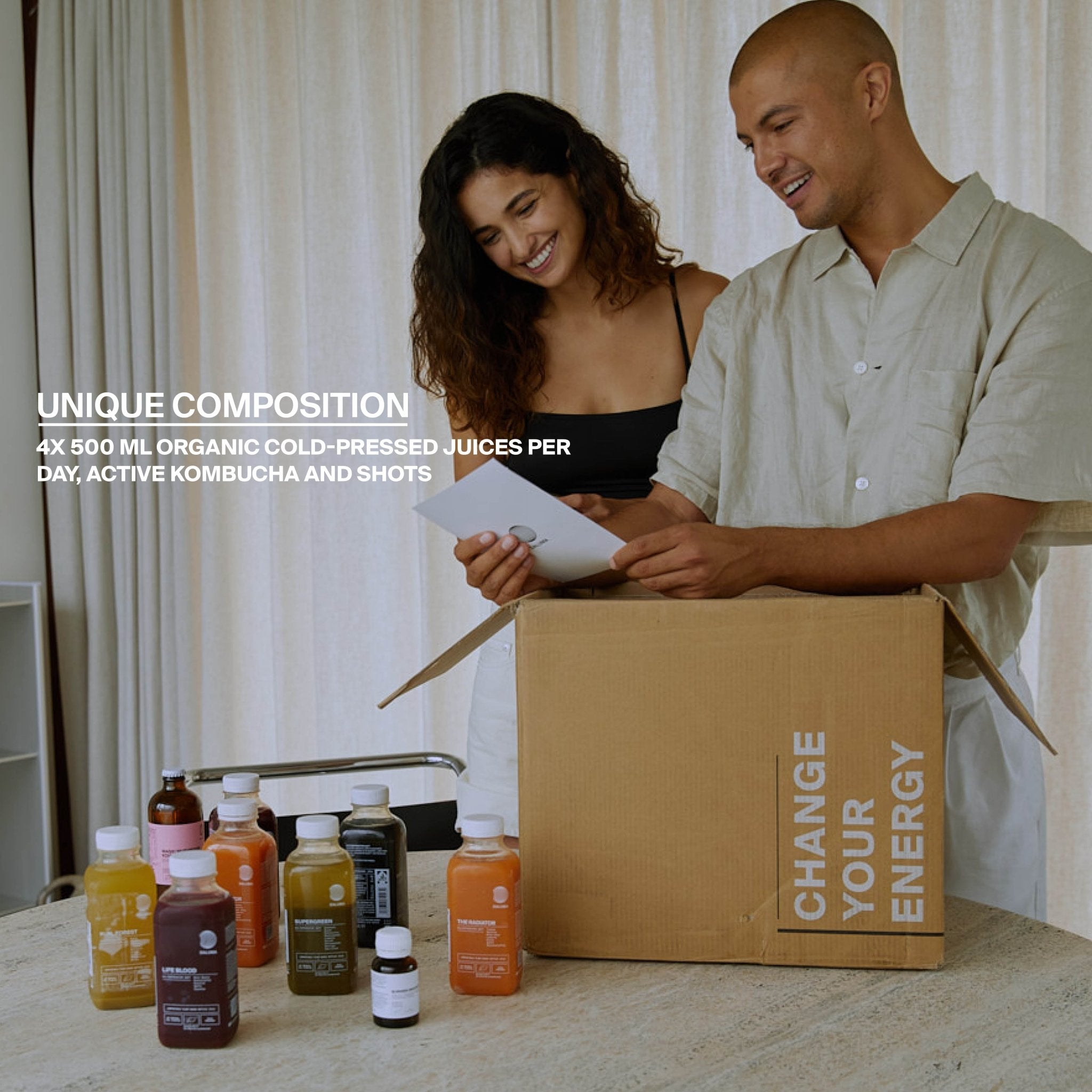 Ein lächelnder Mann und eine lächelnde Frau packen an einem Tisch einen Karton mit der Aufschrift CHANGE YOUR ENERGY aus, umgeben von Flaschen mit DALUMA's BETTER ENERGY SAFTKUR 3 TAGE, bio-kaltgepressten Säften, bunten Säften, unpasteurisiertem Kombucha und Wellness-Drinks.