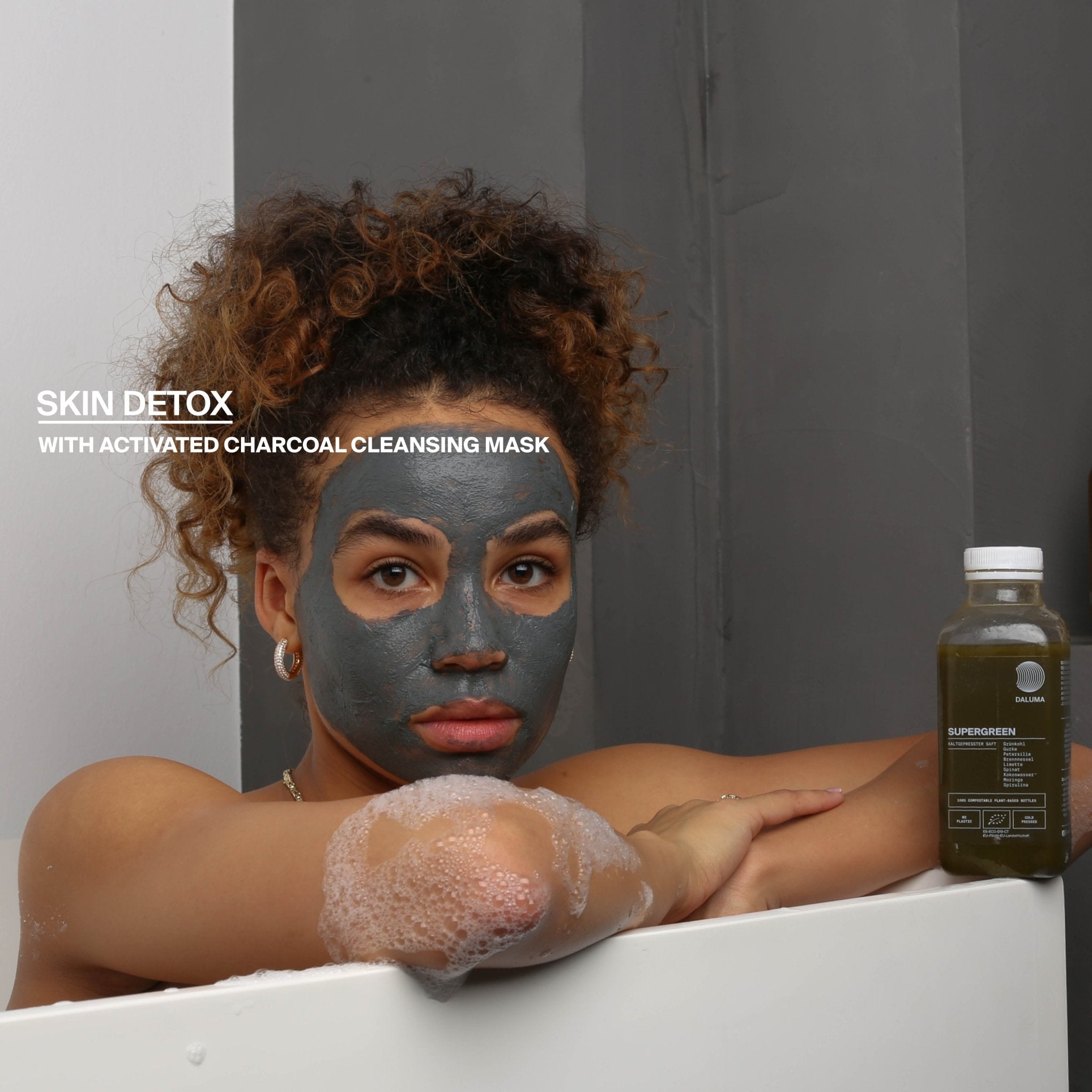 Eine Frau mit lockigem Haar entspannt sich in der Badewanne mit einer Aktivkohlemaske und Seifenblasen. Neben ihr steht eine Flasche DALUMA BETTER ENERGY SAFTKUR 7 TAGE. Text: 