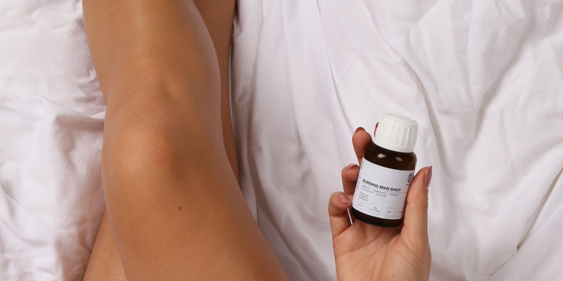 Eine Person mit glatten, nackten Beinen liegt auf einem weißen Laken und hält in einer Hand eine kleine braune Flasche mit weißem Verschluss und Etikett.