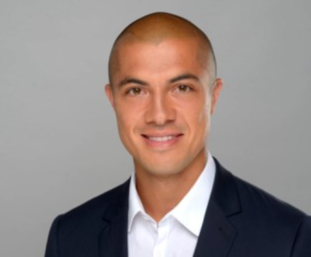 Ein Mann mit rasiertem Kopf lächelt in die Kamera. Er trägt eine dunkle Anzugsjacke über einem weißen Hemd mit Kragen und steht vor einem schlichten hellgrauen Hintergrund.