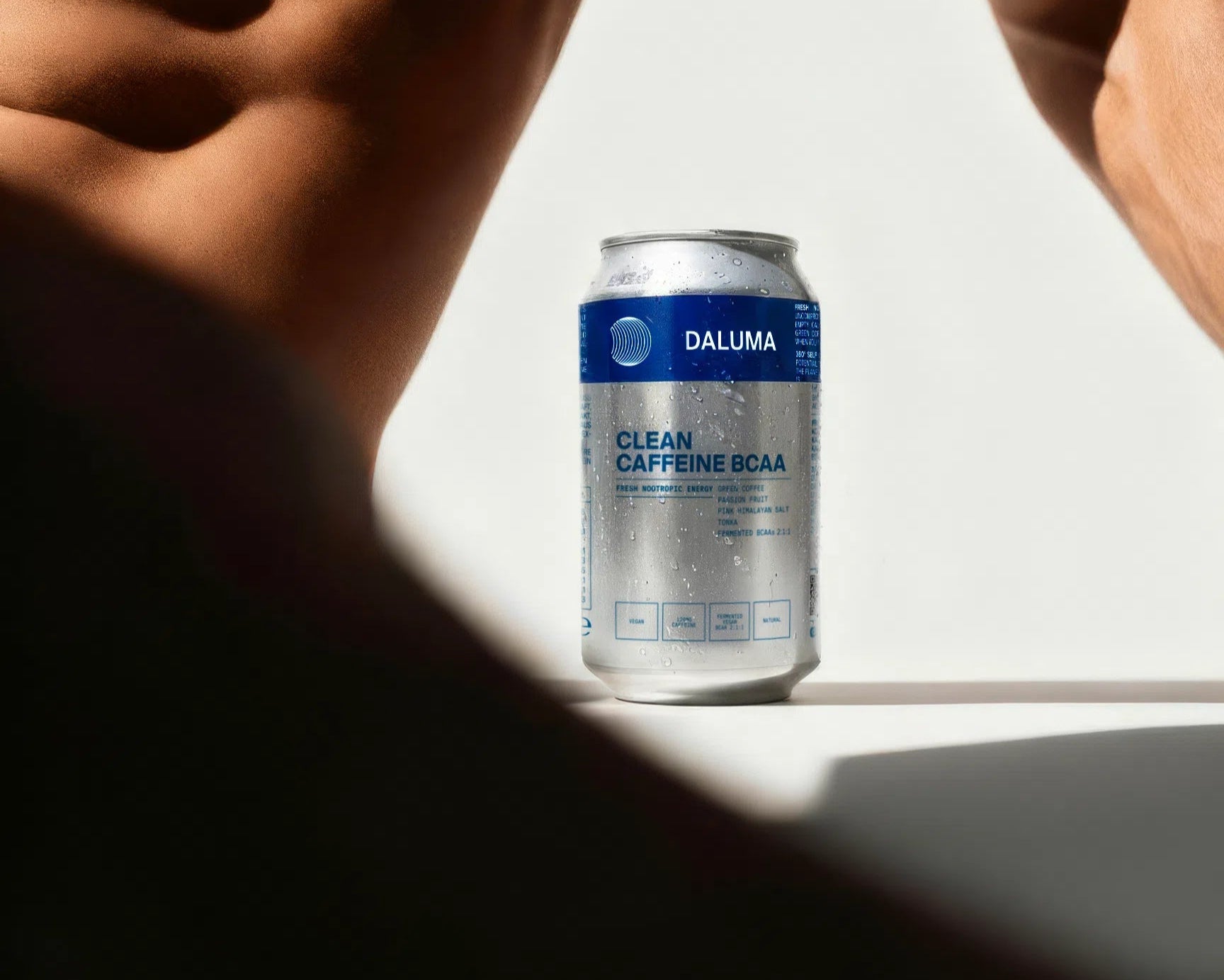 Eine Dose DALUMA CLEAN BCAA ENERGY DRINK mit veganen BCAAs und Wassertropfen steht auf einer weißen Fläche zwischen zwei unscharfen muskulösen Figuren in weichem Naturlicht.