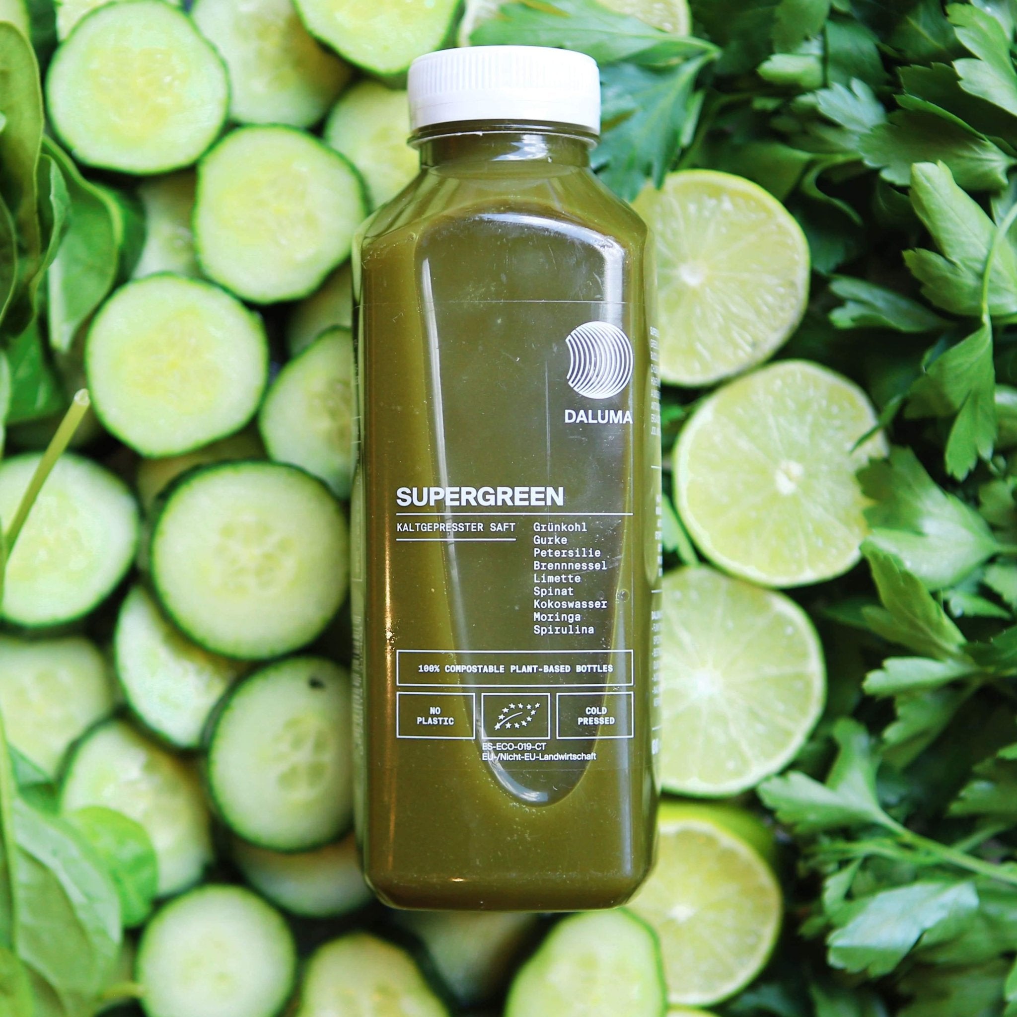 Eine Flasche des DAILY FEEL GOOD JUICE PACKAGE von DALUMA ruht auf Gurken, Limetten und Blattgemüse. Auf dem Etikett stehen nährstoffreicher Spinat und Spirulina. Der Saft ist pflanzlich, biologisch und ideal für Ihr Daily Feel Good-Abo.