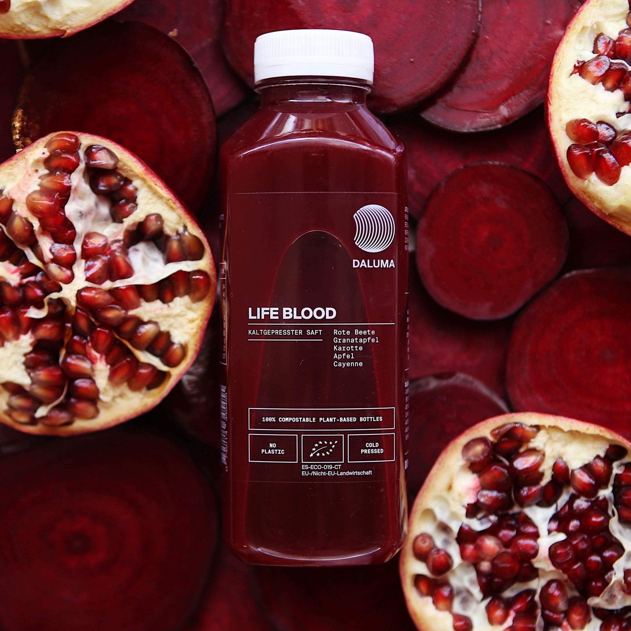 Eine Flasche DALUMA DAILY FEEL GOOD JUICE PACKAGE steht auf einer Fläche mit halbierten Granatäpfeln und in Scheiben geschnittener Roter Bete und stellt ihre natürlichen Inhaltsstoffe zur Schau: Rote Bete, Granatapfel, Karotte, Apfel und Cayenne.