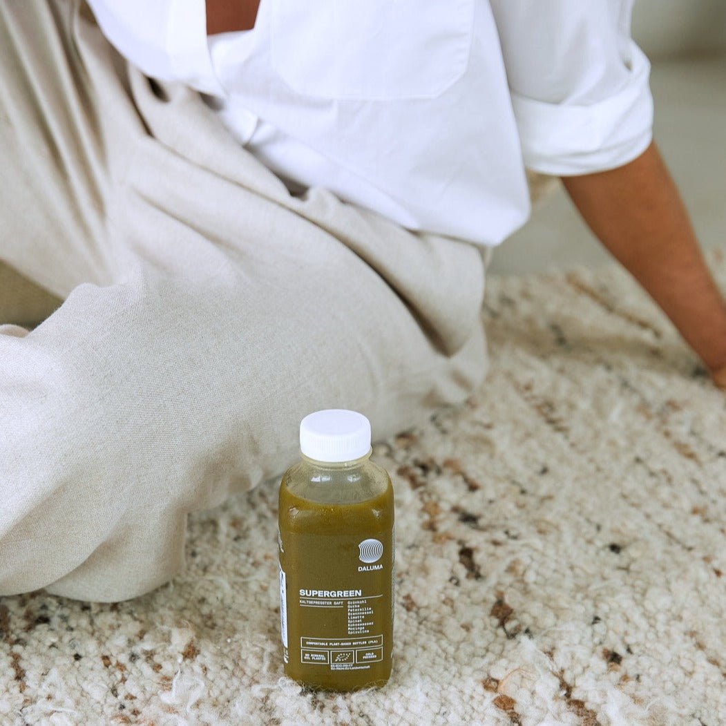 Eine Person in hellbeiger Hose und weißem Hemd sitzt auf einem strukturierten beigen Teppich neben einer durchsichtigen Plastikflasche mit dem DAILY FEEL GOOD JUICE PACKAGE von DALUMA.