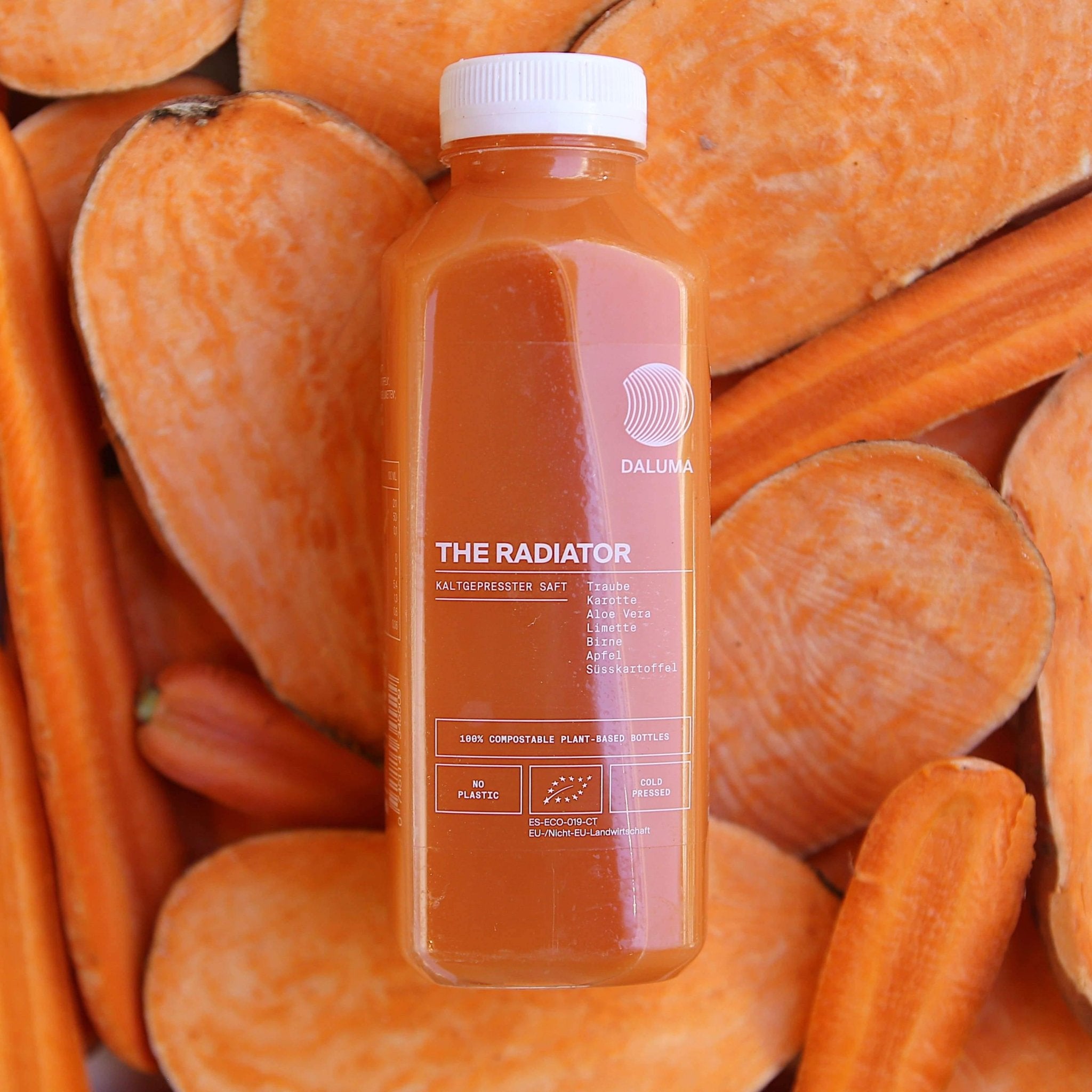 Eine Flasche mit der Aufschrift DAILY FEEL GOOD JUICE PACKAGE von DALUMA, gefüllt mit leuchtendem Orangensaft, steht auf halbierten und in Scheiben geschnittenen Süßkartoffeln - farblich perfekt auf das Gemüse abgestimmt und ideal für Ihr Daily Feel Good-Abo.
