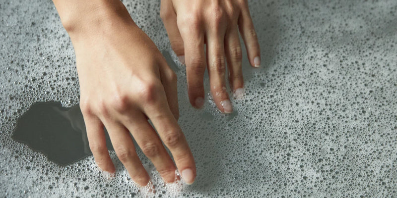 Zwei Hände berühren sanft die Oberfläche von Seifenwasser, wobei Blasen und Schaum um die Finger herum sichtbar sind.