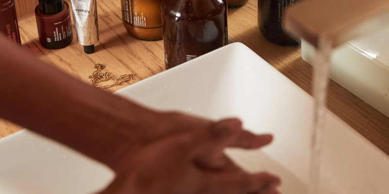 In einem weißen Waschbecken werden die Hände unter fließendem Wasser gewaschen, daneben stehen auf einer hölzernen Arbeitsplatte verschiedene Flaschen und Tuben für die Hautpflege.