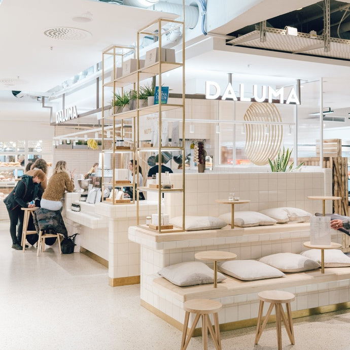 Modernes, minimalistisches Café-Interieur mit weiß gefliesten Oberflächen, Holzakzenten und gepolsterten Sitzgelegenheiten. An der Theke sitzen mehrere Personen. An der Wand hinter der Theke leuchtet das Schild "DALUMA".
