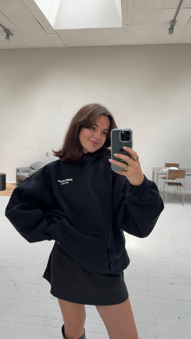 Eine Frau mit schulterlangen braunen Haaren macht ein Selfie im Spiegel in einem hellen, minimalistischen Raum und lächelt dabei in den DALUMA JOSEPH ATELIER X DALUMA REHAB HOODIE. Im Hintergrund sind Möbel und ein großes Fenster zu sehen.